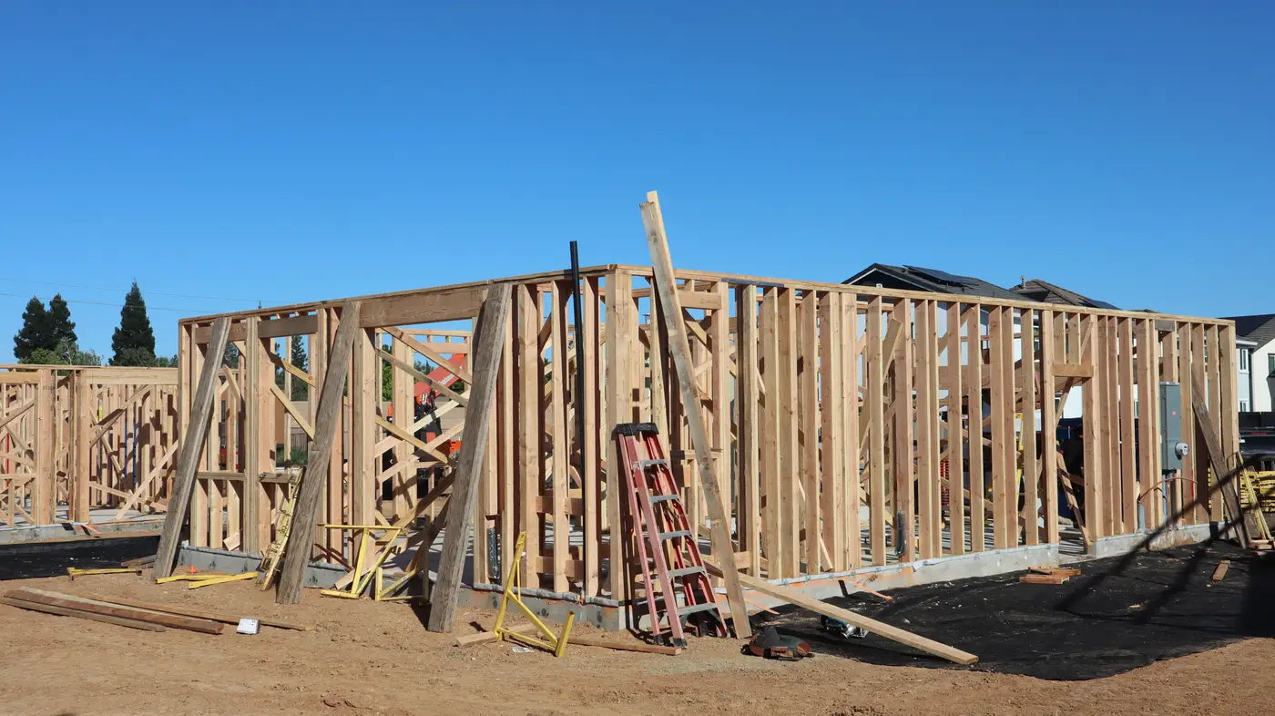 A wood-framed house with bare stud walls standing, no sheathing or roof yet, lumber scattered on the ground.